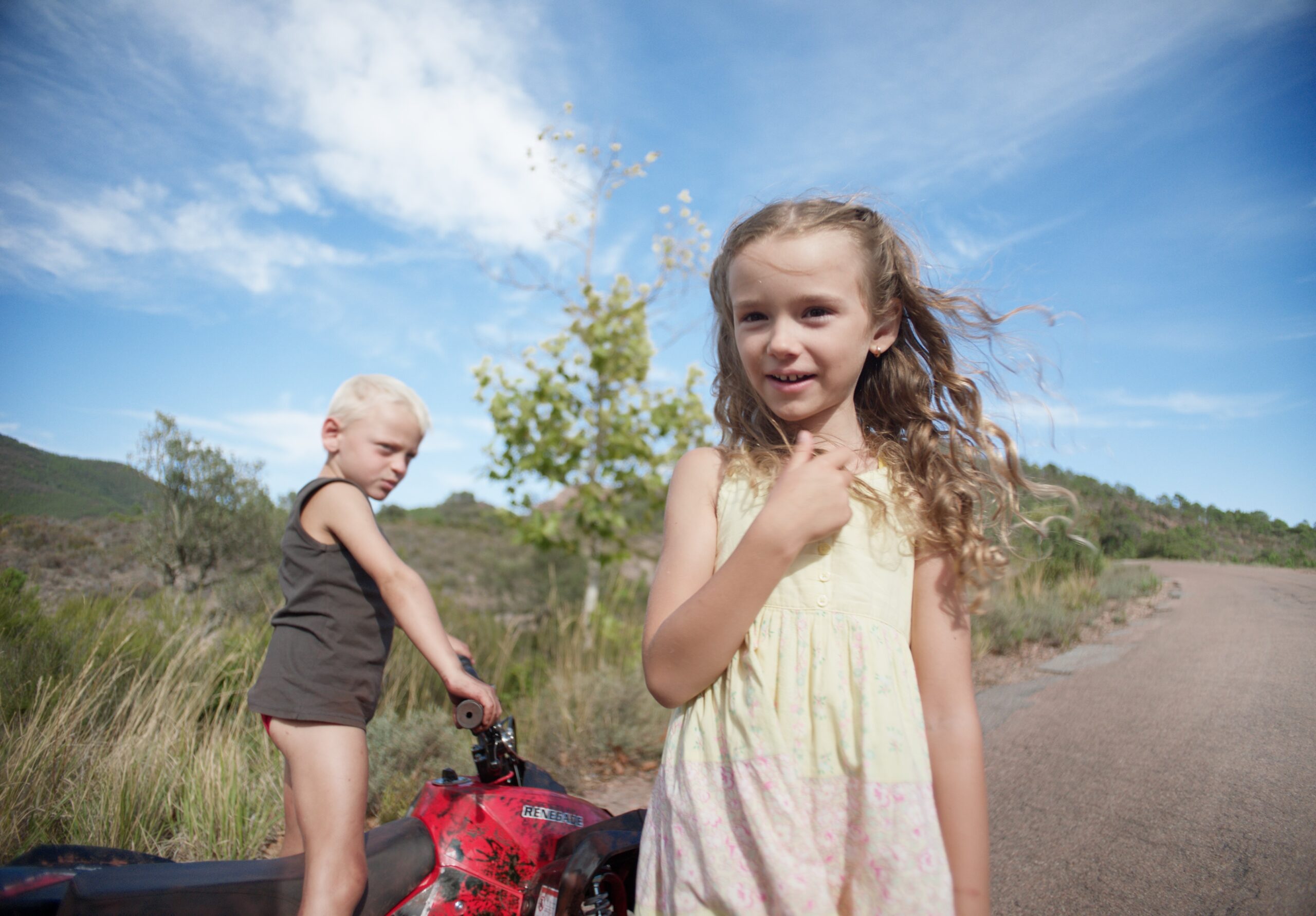 Fotogramma di Red Rocks: un bambino biondo e una bambina accanto a un quad rosso, lungo una strada di campagna
